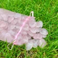 May include: A pink mesh bag filled with heart-shaped rose quartz stones. The stones are a translucent pink color and are scattered on a bed of green grass. The bag has a pink ribbon drawstring.