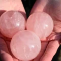 May include: Three polished rose quartz spheres with a translucent, pale pink appearance and natural internal inclusions. Each stone is spherical in shape, featuring a smooth, reflective surface that catches the light, showcasing the mineral's texture.