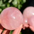May include: A smooth, polished rose quartz sphere with a translucent, pale pink hue. The stone features natural internal fractures and cloudy inclusions throughout its rounded surface, reflecting light with a soft, glossy sheen and a solid, spherical form.