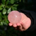 May include: A smooth, polished rose quartz sphere with a translucent, pale pink appearance. The crystal ball features natural internal inclusions and a reflective surface that catches the light, showcasing a soft, rounded shape and consistent color.
