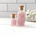 May include: Two glass bottles with cork stoppers filled with pink rose quartz crystals. The bottles are on a white wooden surface.