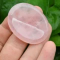 May include: A smooth, oval-shaped rose quartz crystal, polished to a high shine. The crystal is a pale pink color and is translucent. It is being held in a hand.
