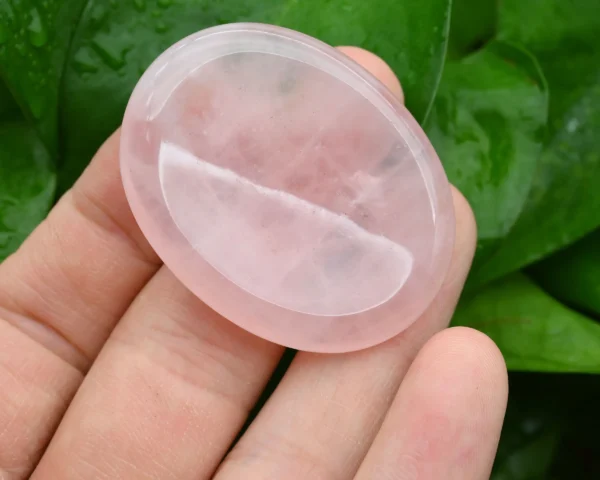 May include: A smooth, oval-shaped rose quartz crystal, polished to a high shine. The crystal is a pale pink color and is translucent. It is being held in a han May include: A smooth, oval-shaped rose quartz crystal, polished to a high shine. The crystal is a pale pink color and is translucent. It is being held in a hand.
