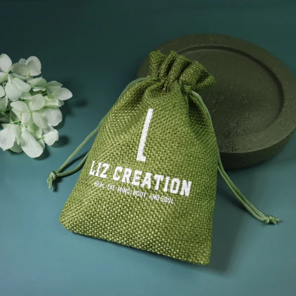 May include: A green burlap drawstring bag with white text that reads "LIZ CREATION" and "HEAL THE MIND, BODY AND SOUL". The bag is tied at the top with a match May include: A green burlap drawstring bag with white text that reads "LIZ CREATION" and "HEAL THE MIND, BODY AND SOUL". The bag is tied at the top with a matching green drawstring. The background is a dark teal color.