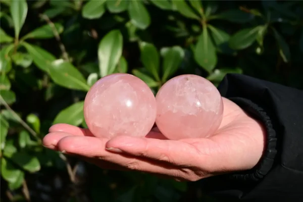 May include: Two smooth, polished rose quartz spheres with a translucent, pale pink hue. The stones exhibit natural internal fractures and cloudy inclusions thr May include: Two smooth, polished rose quartz spheres with a translucent, pale pink hue. The stones exhibit natural internal fractures and cloudy inclusions throughout their rounded surfaces, showcasing the characteristic soft pink color of the mineral.