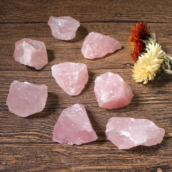 May include: A collection of eight rough-cut rose quartz crystals, each with a soft pink hue. The crystals are scattered on a dark wooden surface, with a few dr May include: A collection of eight rough-cut rose quartz crystals, each with a soft pink hue. The crystals are scattered on a dark wooden surface, with a few dried flowers in the background. The crystals vary in size and shape, showcasing natural textures.