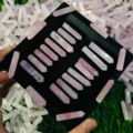 May include: A black velvet box filled with twenty pink crystal wands. The wands are arranged in rows and are all the same size. The box is surrounded by a pile of smaller pink crystals.