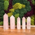 May include: Five polished rose quartz crystal towers arranged in a row on a wooden surface. Each tower features a hexagonal point and a pale pink, translucent appearance. The crystals are numbered one through five in white text at the base.