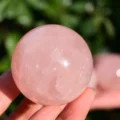 May include: A polished, spherical rose quartz crystal with a translucent, pale pink hue. The stone features natural internal inclusions and a smooth, reflective surface that catches the light, showcasing its soft, consistent pastel coloration.