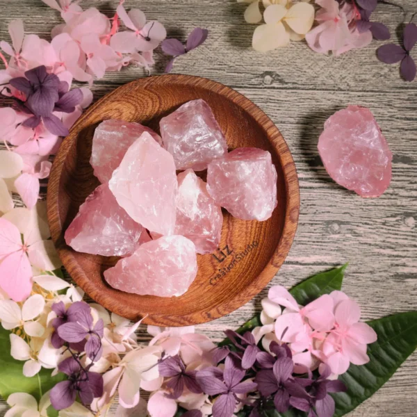 May include: A wooden bowl filled with raw, pink rose quartz crystals. The crystals vary in size and shape, with a translucent appearance. The bowl is surrounde May include: A wooden bowl filled with raw, pink rose quartz crystals. The crystals vary in size and shape, with a translucent appearance. The bowl is surrounded by pink and purple dried flowers, creating a natural, earthy aesthetic.