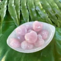 May include: A collection of pink rose quartz heart-shaped crystals in a white bowl. The bowl is sitting on a bed of green leaves.