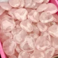 May include: A collection of polished rose quartz heart-shaped stones. The stones are a soft pink color and appear smooth and translucent. The hearts are of varying sizes and are piled together.