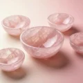 May include: Five rose quartz bowls of varying sizes are arranged on a pink surface. The bowls have a translucent, pink hue with natural veining patterns. The bowls are smooth and polished, reflecting light.