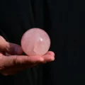May include: A smooth, polished sphere made of translucent pale pink rose quartz. The stone features natural internal inclusions and a glossy surface that reflects light, showcasing a soft, uniform pastel pink color throughout the entire round object.