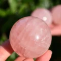 May include: A polished, spherical rose quartz crystal with a translucent, pale pink hue. The surface of the stone displays natural internal fractures and cloudy inclusions, reflecting light with a smooth, rounded finish and a soft, light pink color.
