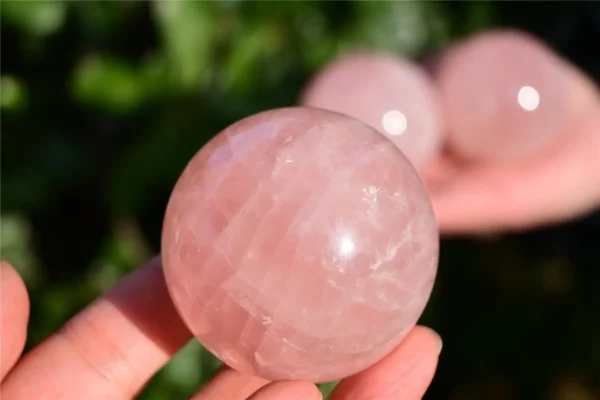 May include: A polished, spherical rose quartz crystal with a translucent, pale pink hue. The surface of the stone displays natural internal fractures and cloud May include: A polished, spherical rose quartz crystal with a translucent, pale pink hue. The surface of the stone displays natural internal fractures and cloudy inclusions, reflecting light with a smooth, rounded finish and a soft, light pink color.