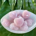 May include: A bowl filled with eight pink heart-shaped rose quartz crystals. The bowl is white and sits on a green leafy surface.