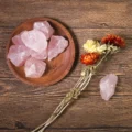May include: A collection of raw, pink rose quartz crystals displayed in a wooden bowl and scattered on a wooden surface. Dried flowers in shades of orange and yellow are also present, alongside a single rose quartz piece.