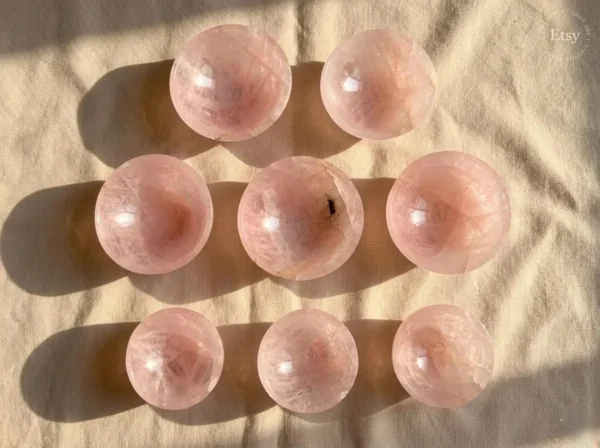 May include: Nine polished rose quartz spheres, each with a translucent pink hue, are arranged on a beige fabric. The spheres vary slightly in color and interna May include: Nine polished rose quartz spheres, each with a translucent pink hue, are arranged on a beige fabric. The spheres vary slightly in color and internal patterns, with shadows cast by the sunlight.