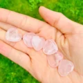 May include: A hand holding several heart-shaped rose quartz stones. The stones are a soft pink color and appear smooth and polished. The background is a blurred green.
