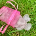 May include: A pink mesh drawstring bag spilling heart-shaped rose quartz stones onto green grass. The stones are a pale pink color and appear smooth and polished. The bag is a vibrant pink.