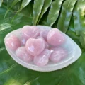 May include: A white bowl filled with pink heart-shaped rose quartz crystals. The bowl is sitting on a green leafy surface.
