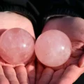 May include: Two polished rose quartz spheres with a translucent, pale pink appearance. Each stone features a smooth, rounded surface with natural internal inclusions and subtle variations in color, reflecting light across their curved forms.