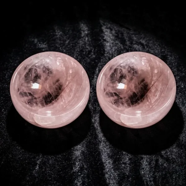 May include: Two polished rose quartz spheres. The spheres are a soft pink color with internal veining and a smooth, reflective surface. The spheres are display May include: Two polished rose quartz spheres. The spheres are a soft pink color with internal veining and a smooth, reflective surface. The spheres are displayed on a dark, textured background.