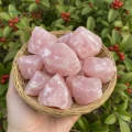 May include: A woven basket filled with raw, light pink rose quartz crystals. The crystals have a rough, natural texture and varying shapes. The basket is held against a backdrop of green leaves and red berries.