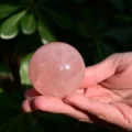 May include: A polished, spherical rose quartz crystal with a translucent, pale pink hue. The stone features natural internal inclusions and a smooth, reflective surface that catches the light, showcasing its soft, semi-opaque, and crystalline texture.